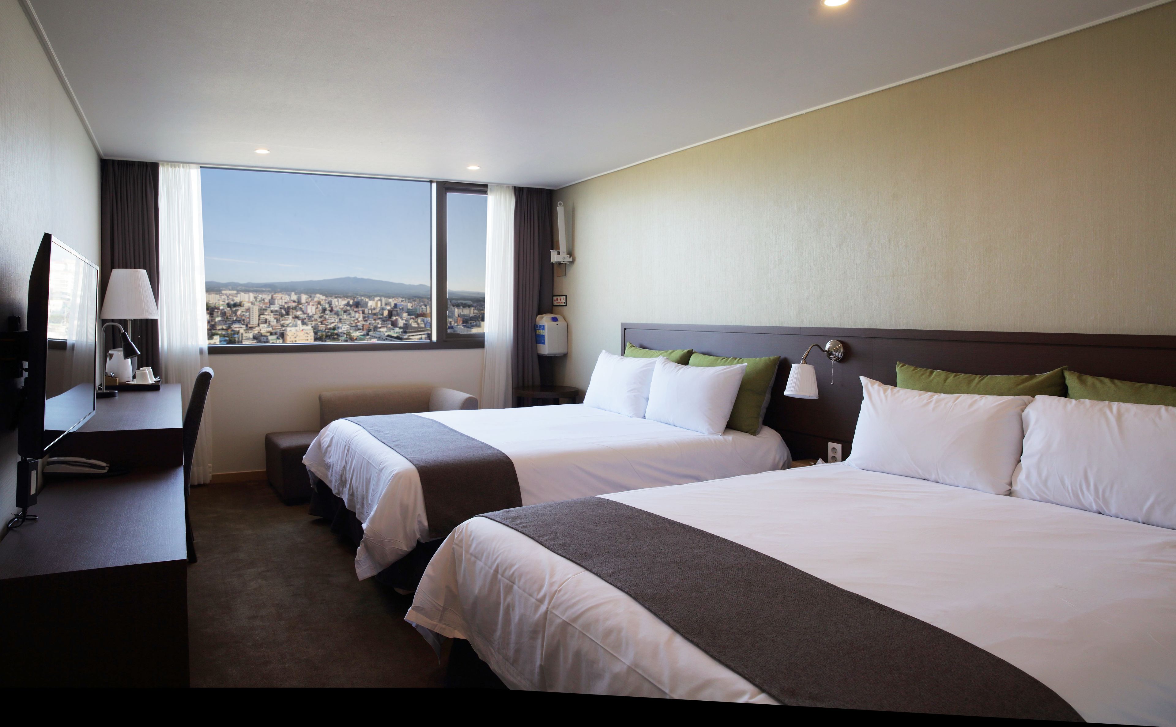 A hotel room with two beds, a desk, a chair, a lamp, and a view of a cityscape.