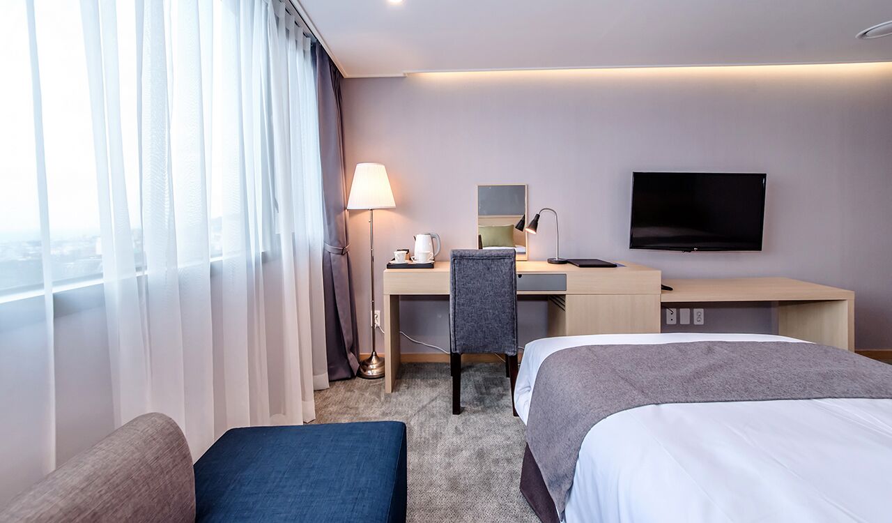 A hotel room with a bed, a desk with a chair, a TV, and a window with curtains.