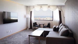 Una camera d'albergo con televisione a schermo piatto, un divano, due poltrone, un tavolino e vista sulla città attraverso ampie finestre.