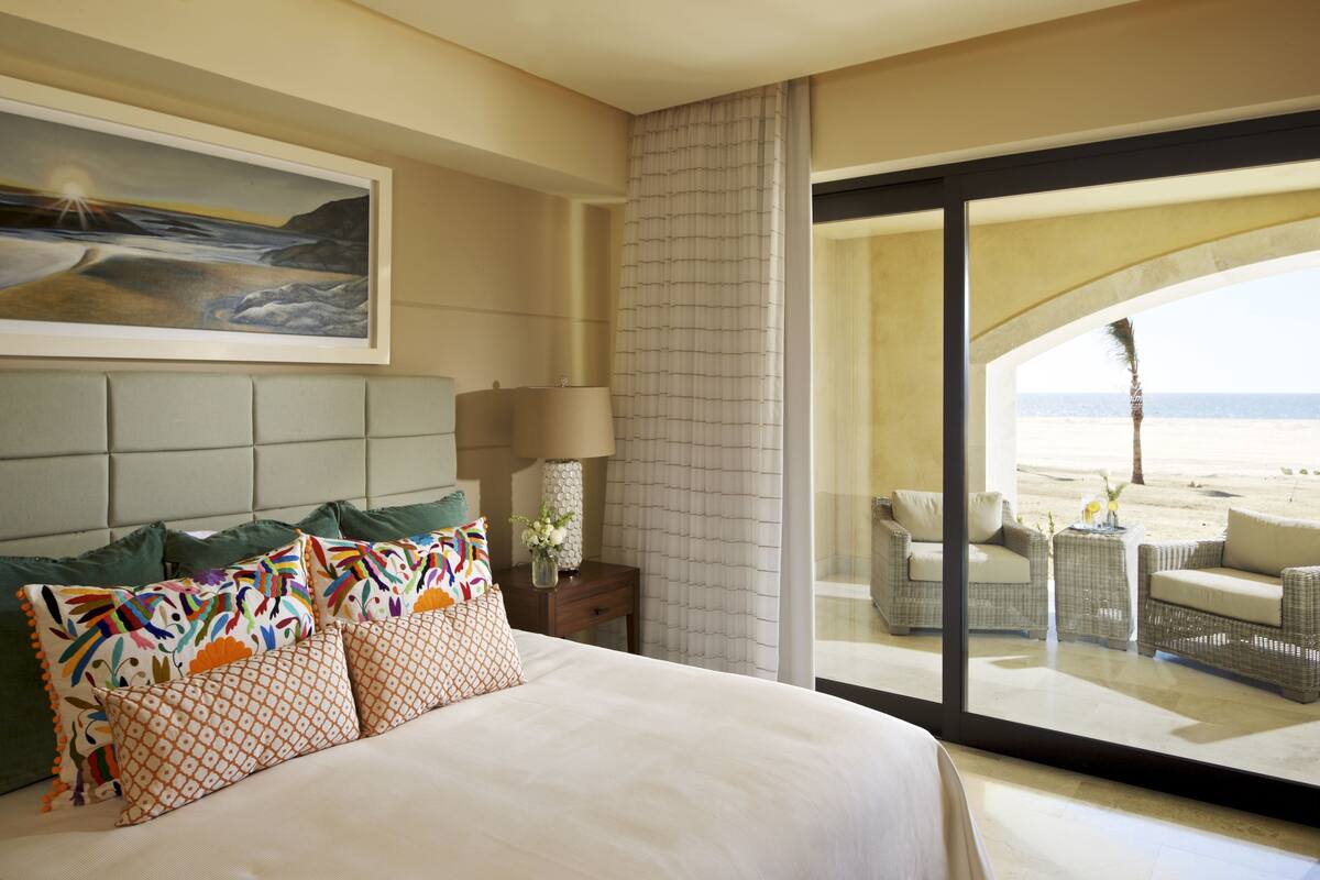 A hotel room with a large bed, a painting on the wall, a view of the beach through a glass door, and a balcony with wicker furniture.