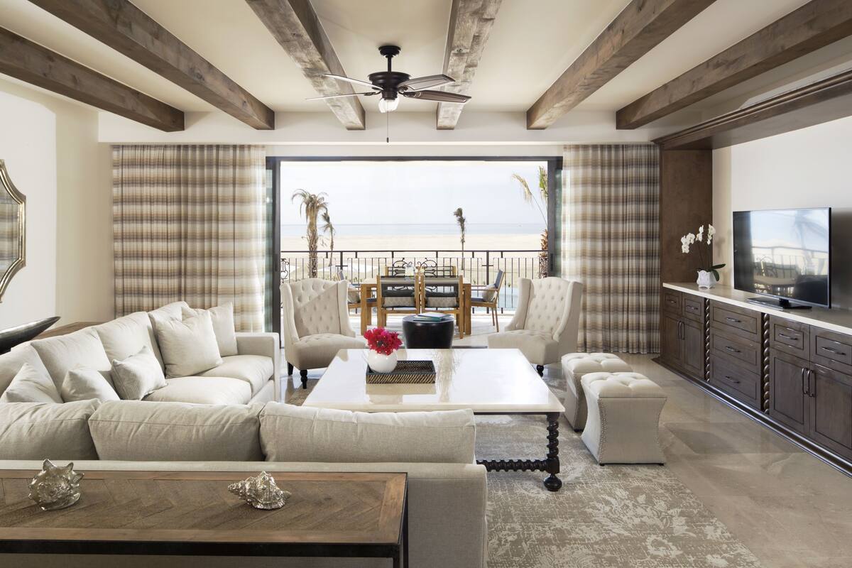 A spacious living room with a large sofa, a coffee table, and a television. There is a balcony with a view of the beach.