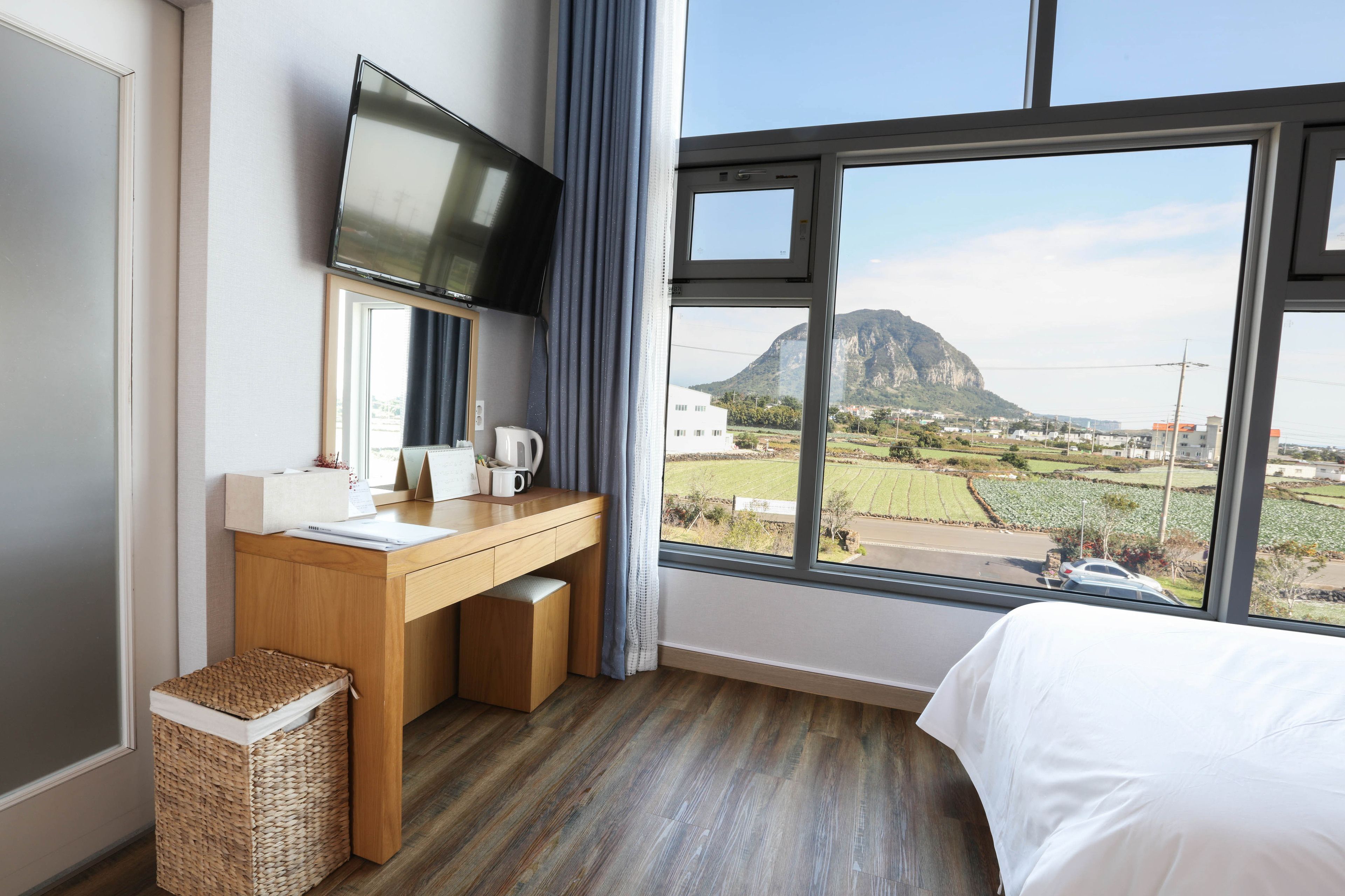 A hotel room with a wooden desk, a flat-screen TV, a bed, and a large window offering a view of a mountain and fields.