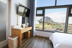 Habitación de hotel con un escritorio de madera, un televisor de pantalla plana, una cama y un amplio ventanal con vistas a una montaña y campos.