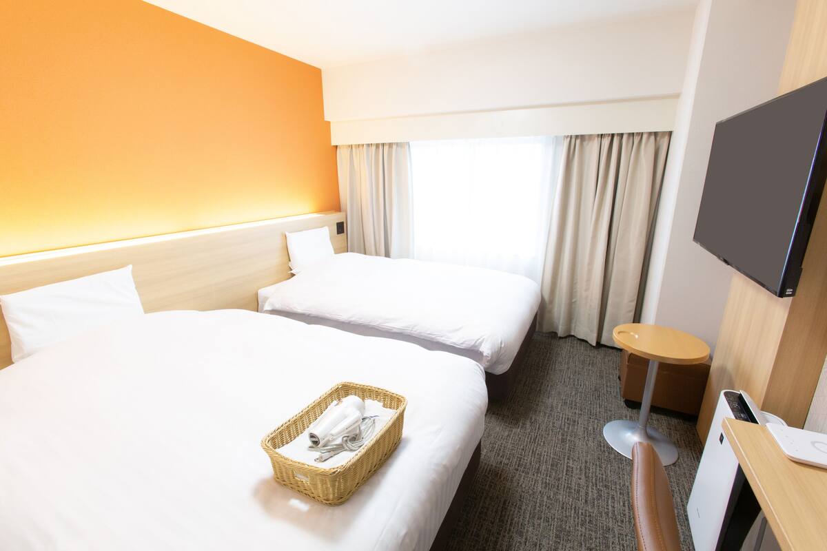 A hotel room with two beds, a TV, a small table, and a window with curtains.