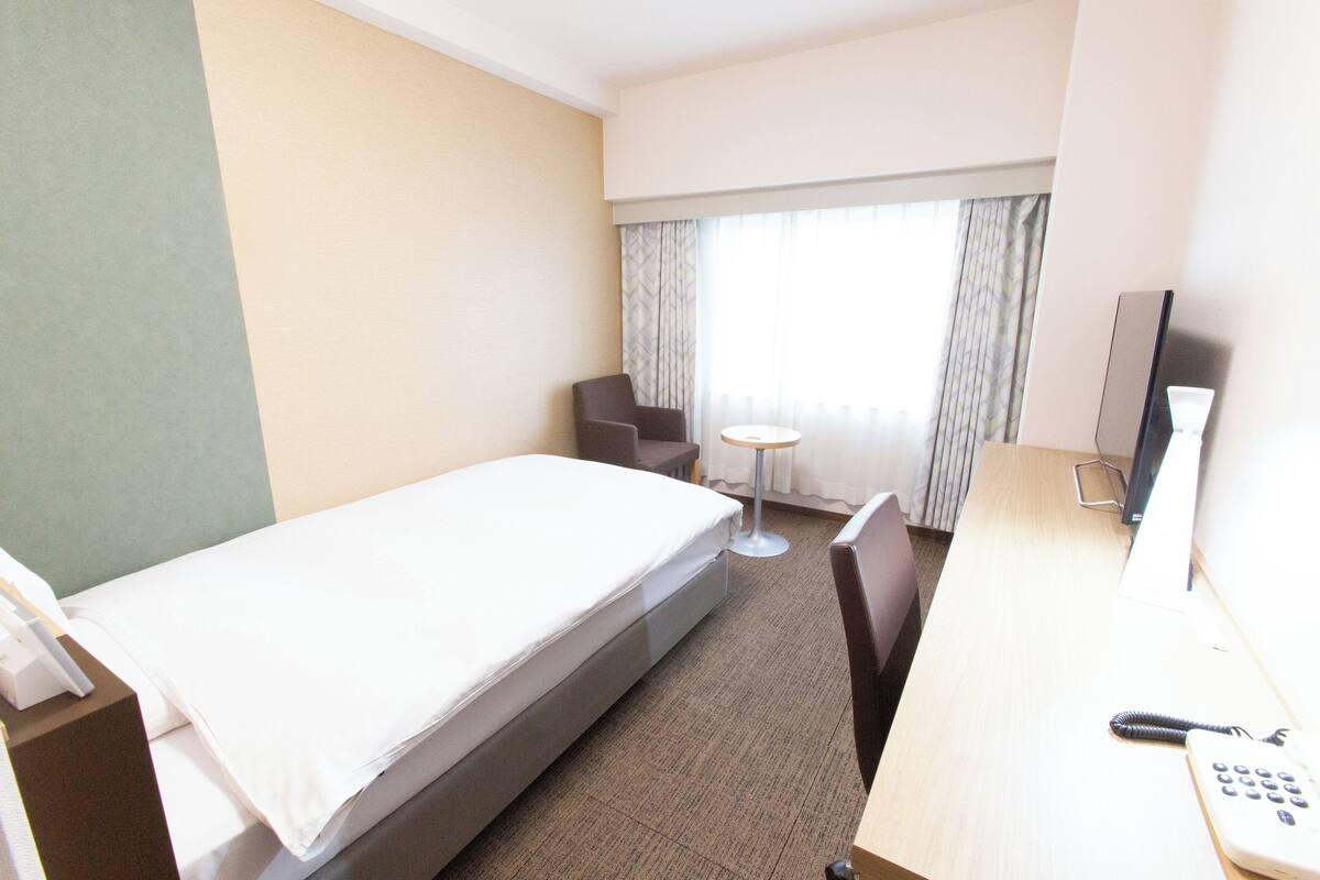 A hotel room with a single bed, a desk with a telephone, a chair, a small round table, and a window with curtains.