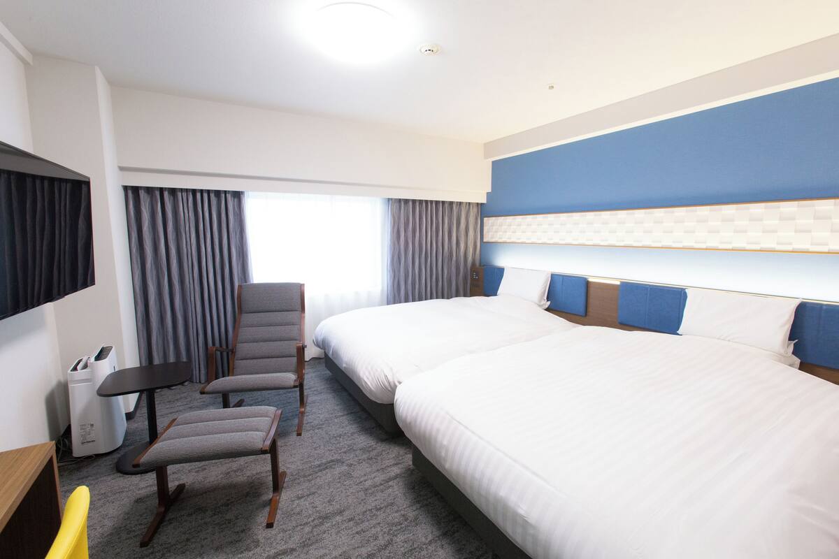 A hotel room with two beds, a desk, a chair, and a TV.