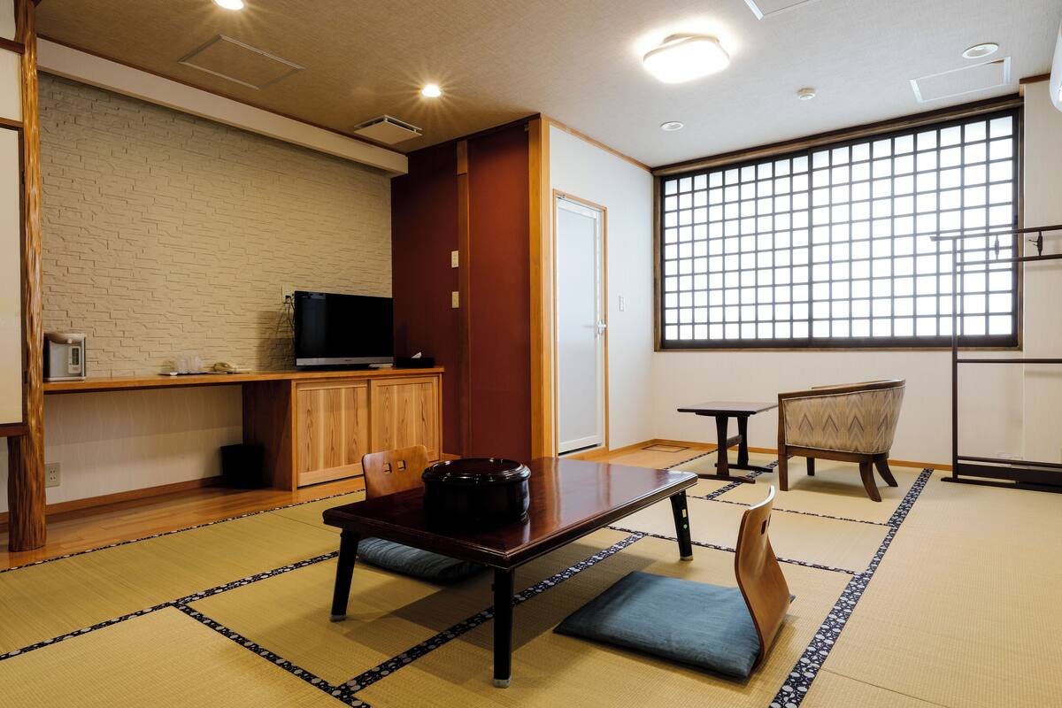 Une pièce de style japonais traditionnel, avec un sol recouvert de tatamis, une petite table sur laquelle se trouve un récipient noir, un téléviseur posé sur un meuble en bois et une grande fenêtre à carreaux.