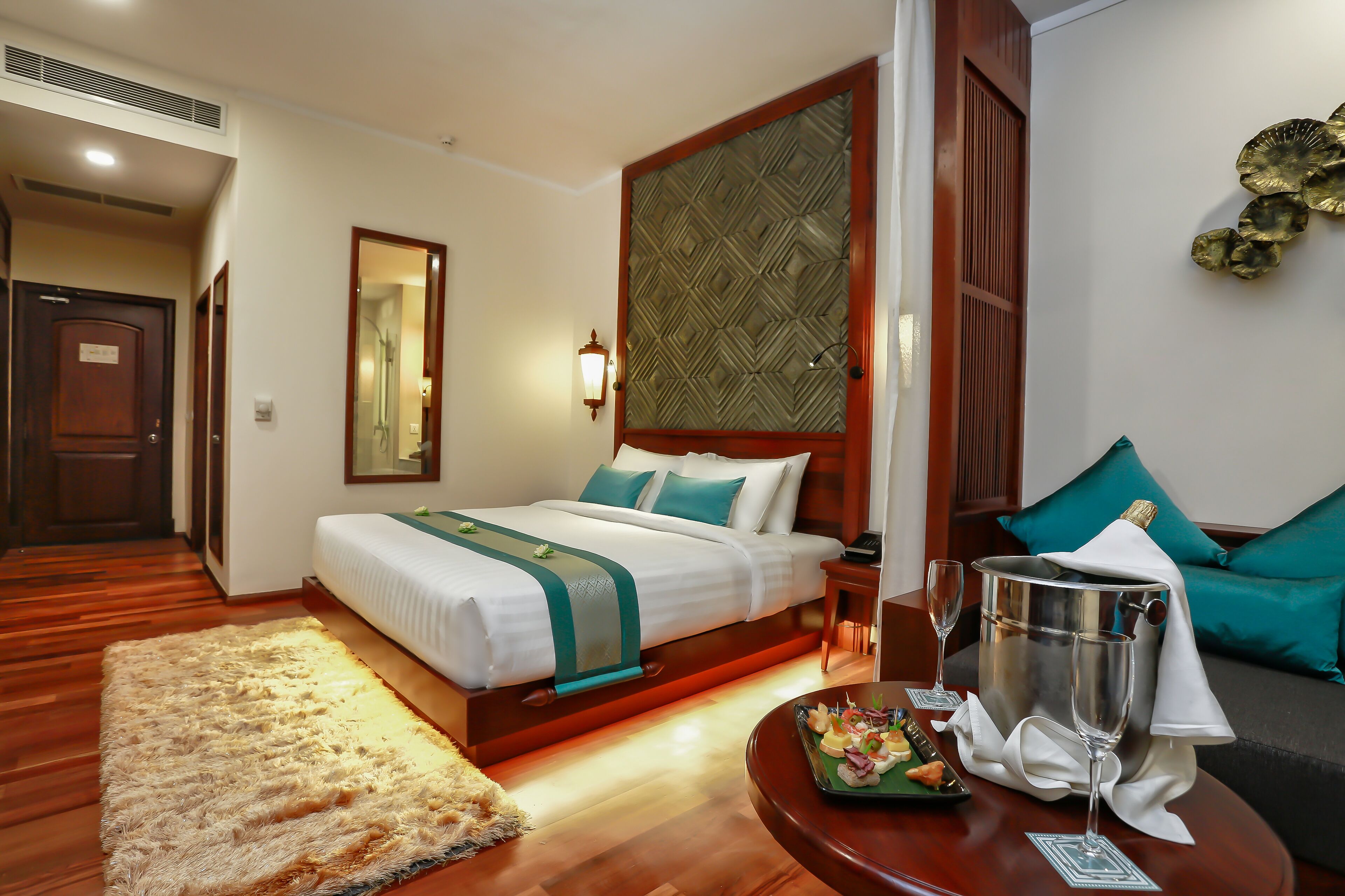 Una habitación de hotel con una cama, una mesita de noche, una mesa redonda con una bandeja de comida y vasos, una alfombra mullida y un adorno en la pared.