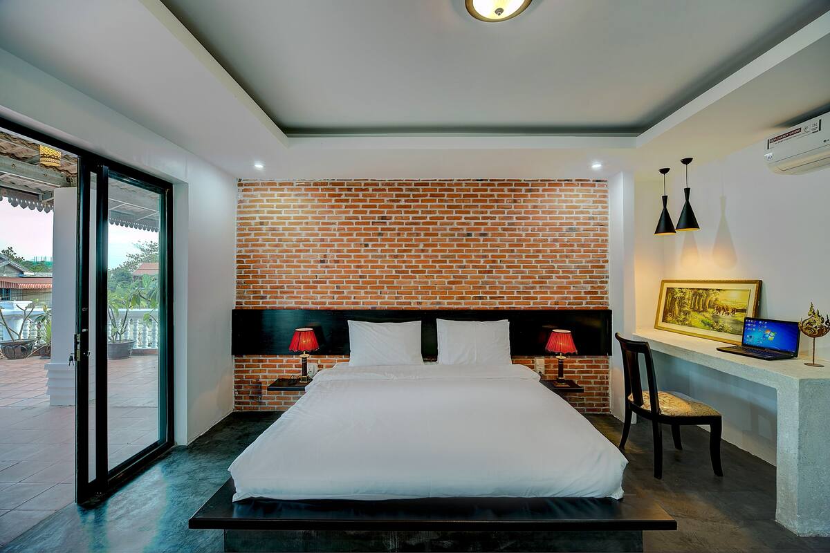 A modern bedroom with a brick-patterned headboard, a large bed, a desk with a laptop, and a red lamp.