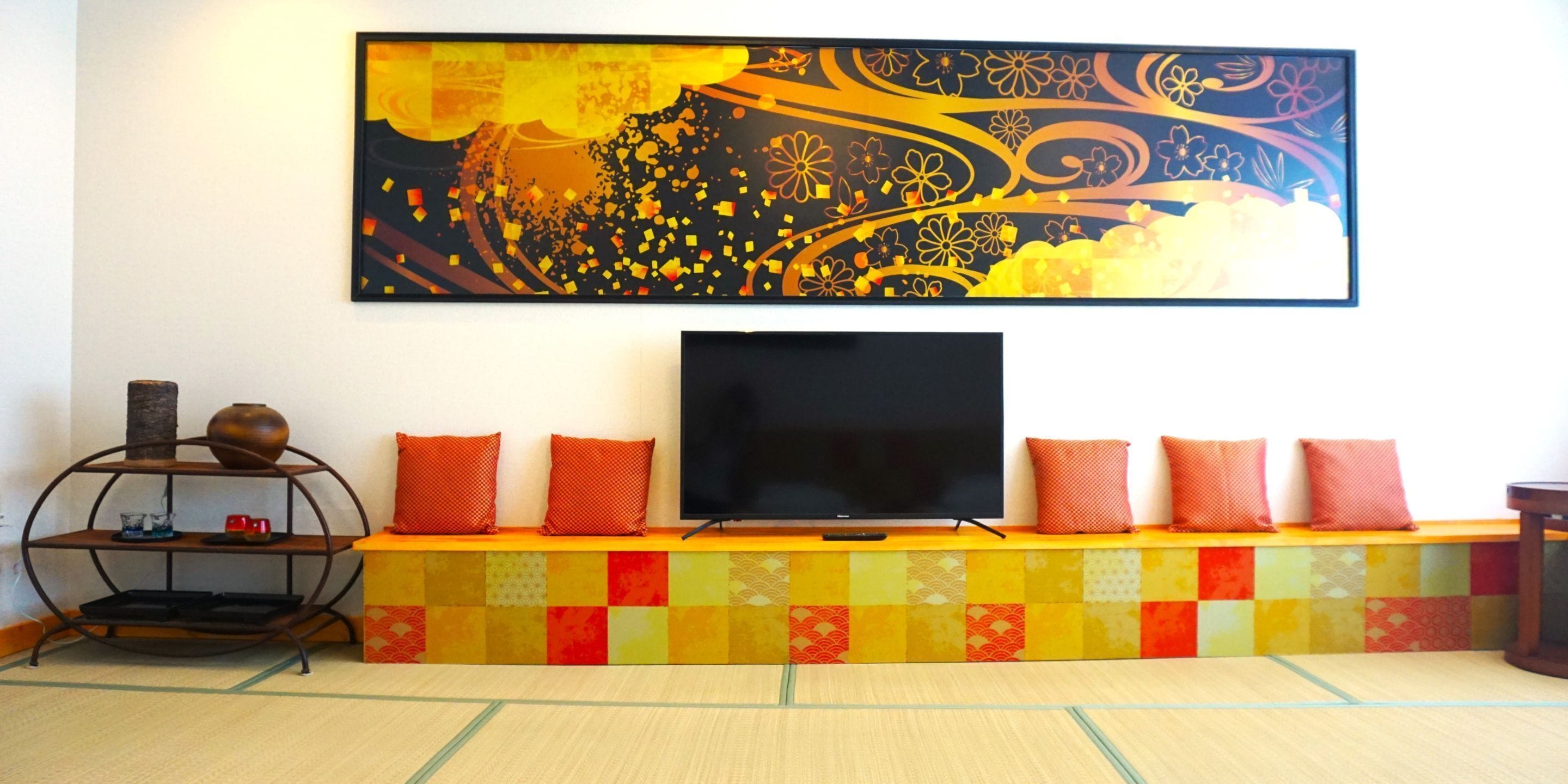 A living room with a flat-screen TV on a colorful bench, a decorative shelf with a vase, and a large abstract painting on the wall.