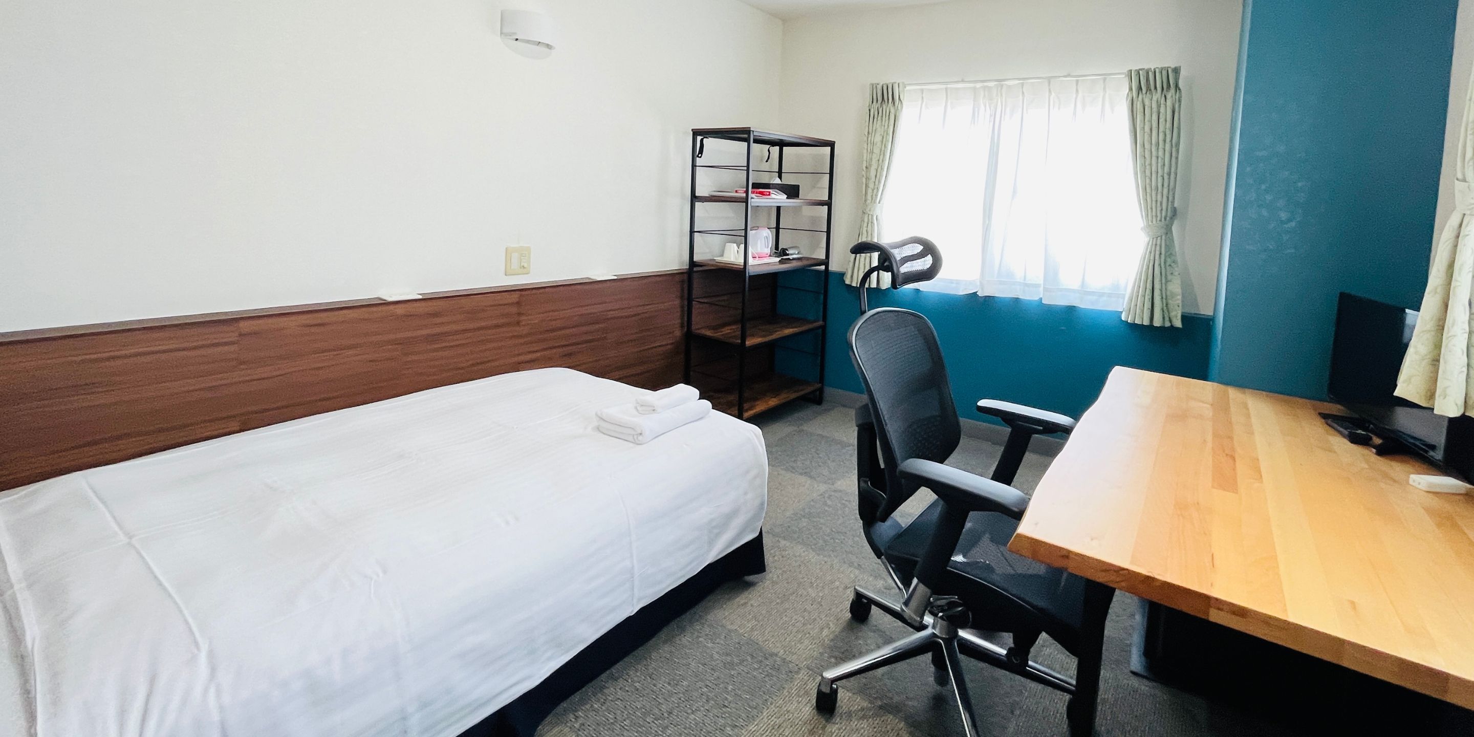 A hotel room with a single bed, a desk with a chair, a bookshelf, and a television.