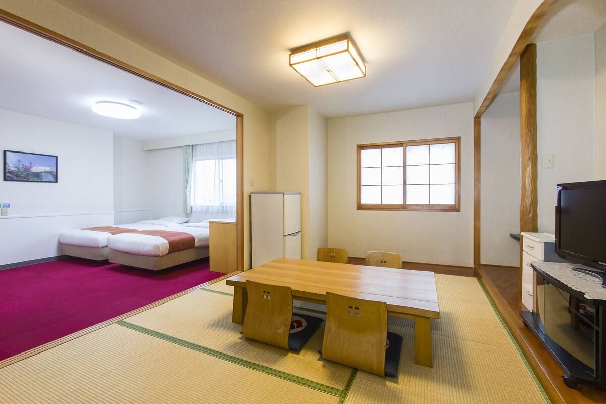 A room with a wooden table, chairs, a bed, a television, and a window.