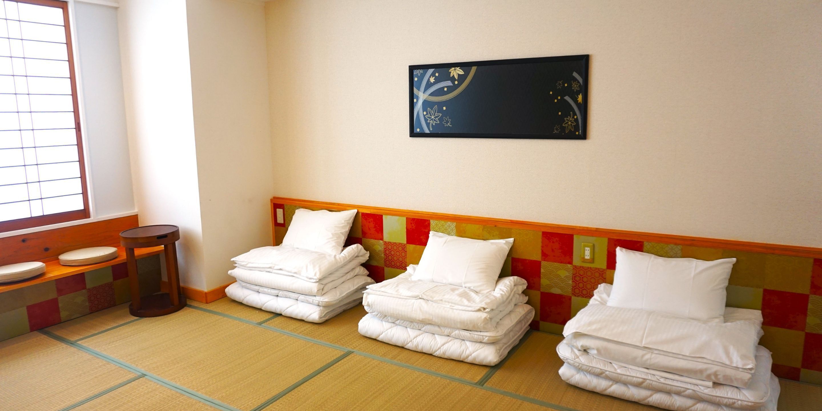 Habitación de hotel con suelo de tatami, cama con ropa blanca, una mesita redonda y una obra de arte colgada en la pared.
