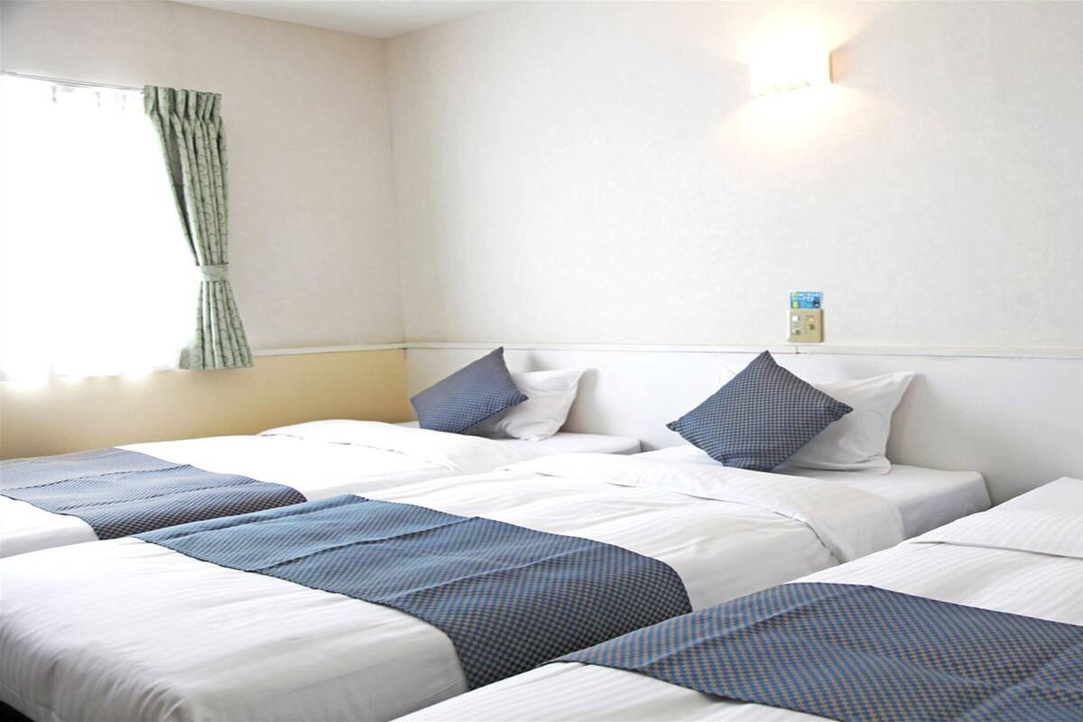 A hotel room with three beds, each with white bedding and blue accents. There is a window with curtains, a wall-mounted light, and a small box on the wall.