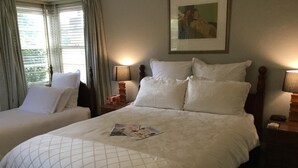 A hotel room with two beds, each with white bedding and a quilted pattern. There are two bedside tables with lamps, a framed picture on the wall, and a window with curtains. - The Evergreen B&B (Braddon)