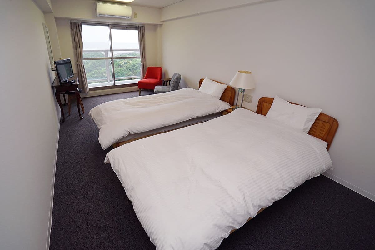 A hotel room with two beds, a desk with a computer, a chair, and a window with a view.