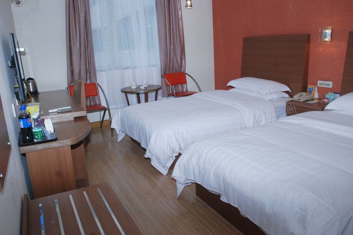 Ein Hotelzimmer mit zwei Betten, einem Schreibtisch mit Computer, einem Stuhl, einem kleinen Tisch mit Telefon und Tasse sowie einem Fenster mit Vorhängen.