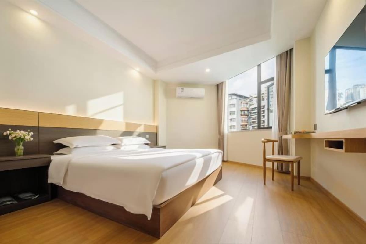 A modern hotel room with a large bed, a bedside table, a chair, and a window with a city view.