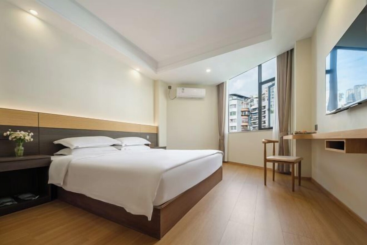 A hotel room with a bed, bedside table, chair, and large window with a city view.