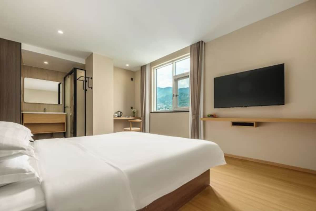 A hotel room with a bed, a TV mounted on the wall, a window with a view, and a bathroom with a mirror and sink.