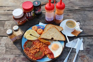 Un plato de comida para el desayuno con tostadas, huevos, frijoles, tomates y tocino, junto con salsas y una taza de café sobre una mesa de madera.