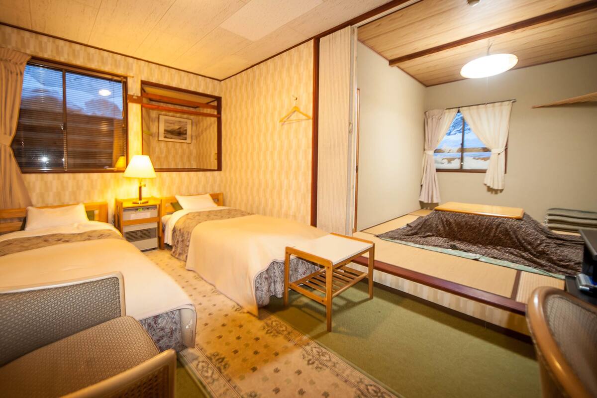 A hotel room with two beds, a small table, a chair, a desk, and a window with curtains.