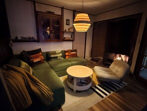 Een gezellige woonkamer met een hoekbank, een ronde salontafel, een witte fauteuil en een open haard.