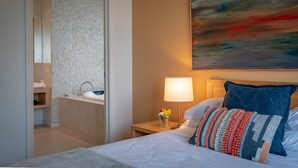 A hotel room with a bed, a nightstand with a lamp, a painting on the wall, and a bathroom visible through an open door. - Queensberry Springs Villa 1 (Daylesford)
