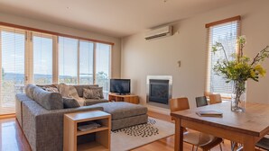 A modern living room with a grey sofa, a flat-screen TV, a fireplace, and a dining table with chairs. - Queensberry Springs Villa 1 (Daylesford)