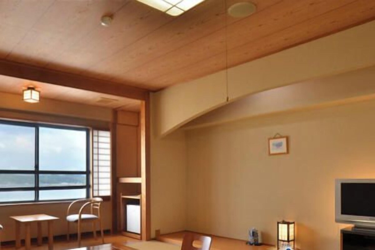 A room with a wooden ceiling, a large window with a view, a television, and a table with chairs.