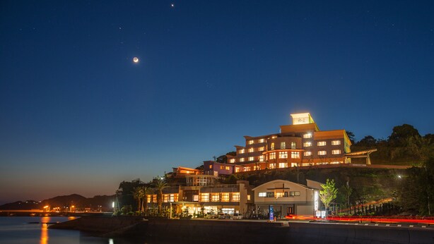 A multi-story hotel with a prominent rooftop structure, situated by a waterfront with a calm body of water reflecting lights. - Hotel Shouryuen Kaisei (Kamiamakusa)