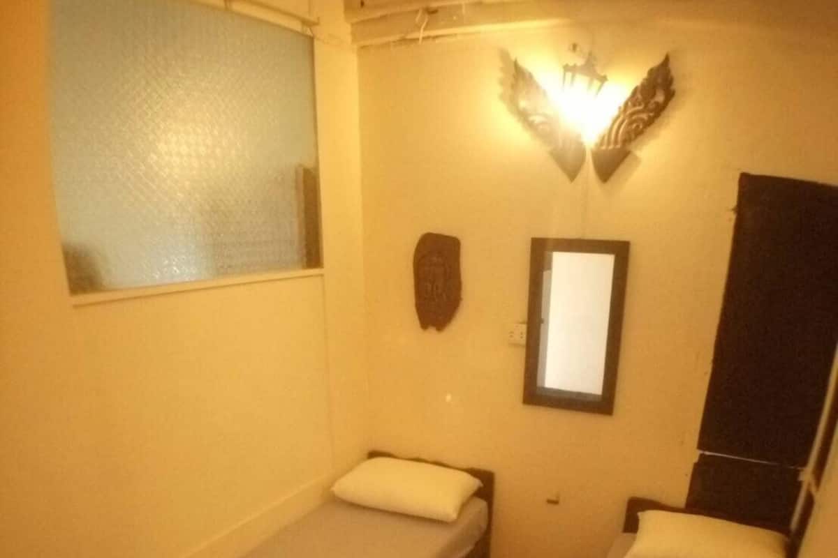 Ein kleiner, schwach beleuchteter Raum mit zwei Einzelbetten, jeweils mit Kissen und Decke. An der linken Wand befindet sich ein Fenster, über den Betten eine Wandleuchte und eine kleine Holzdeko an der Wand.