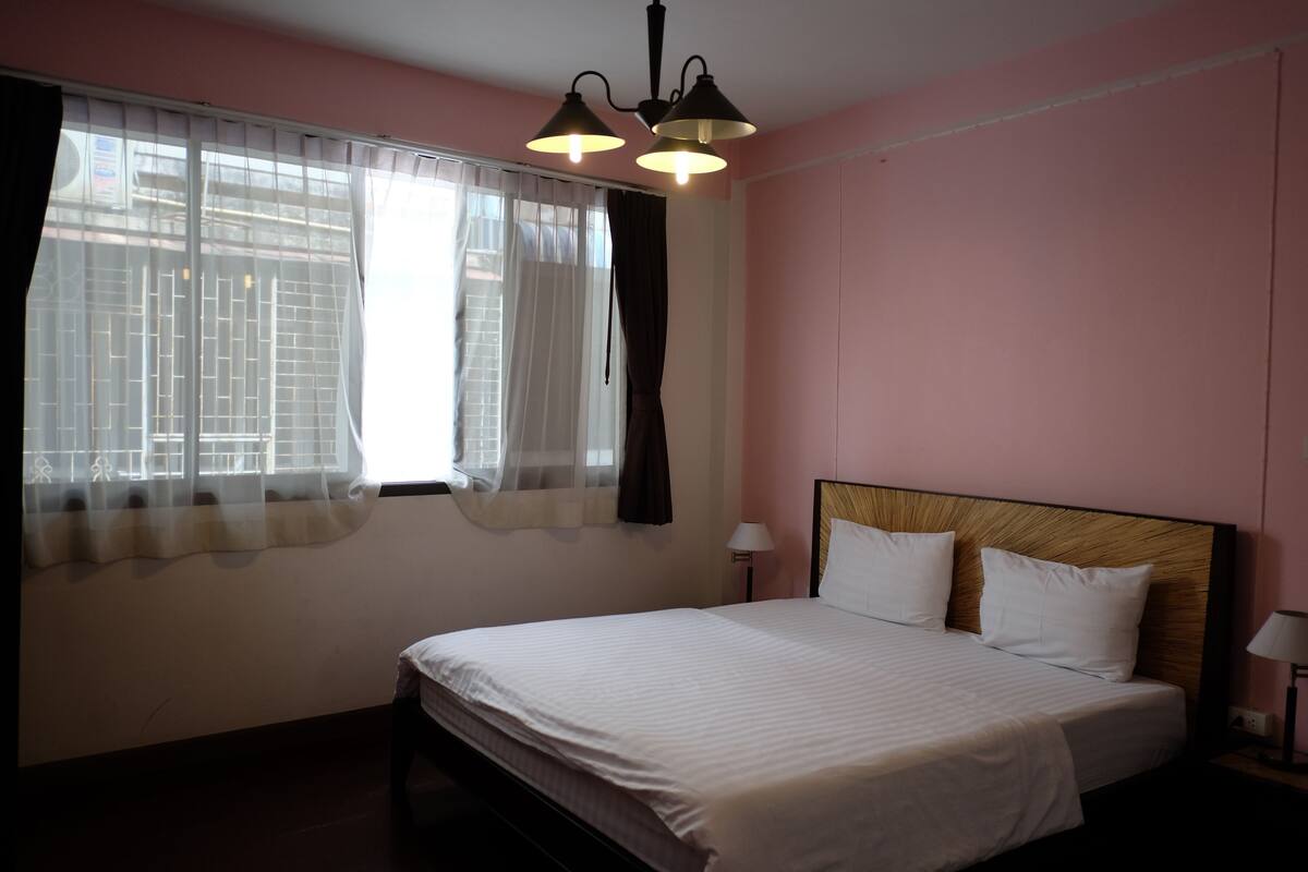 Une chambre à coucher avec un lit, deux lampes de chevet, une fenêtre avec des rideaux légers et un luminaire au plafond.