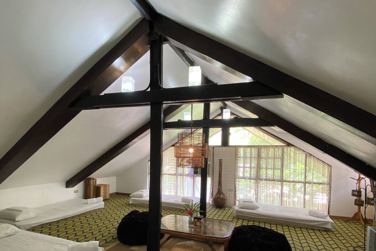 A cozy attic room with a sloped ceiling, two beds, a small table, and a large window.