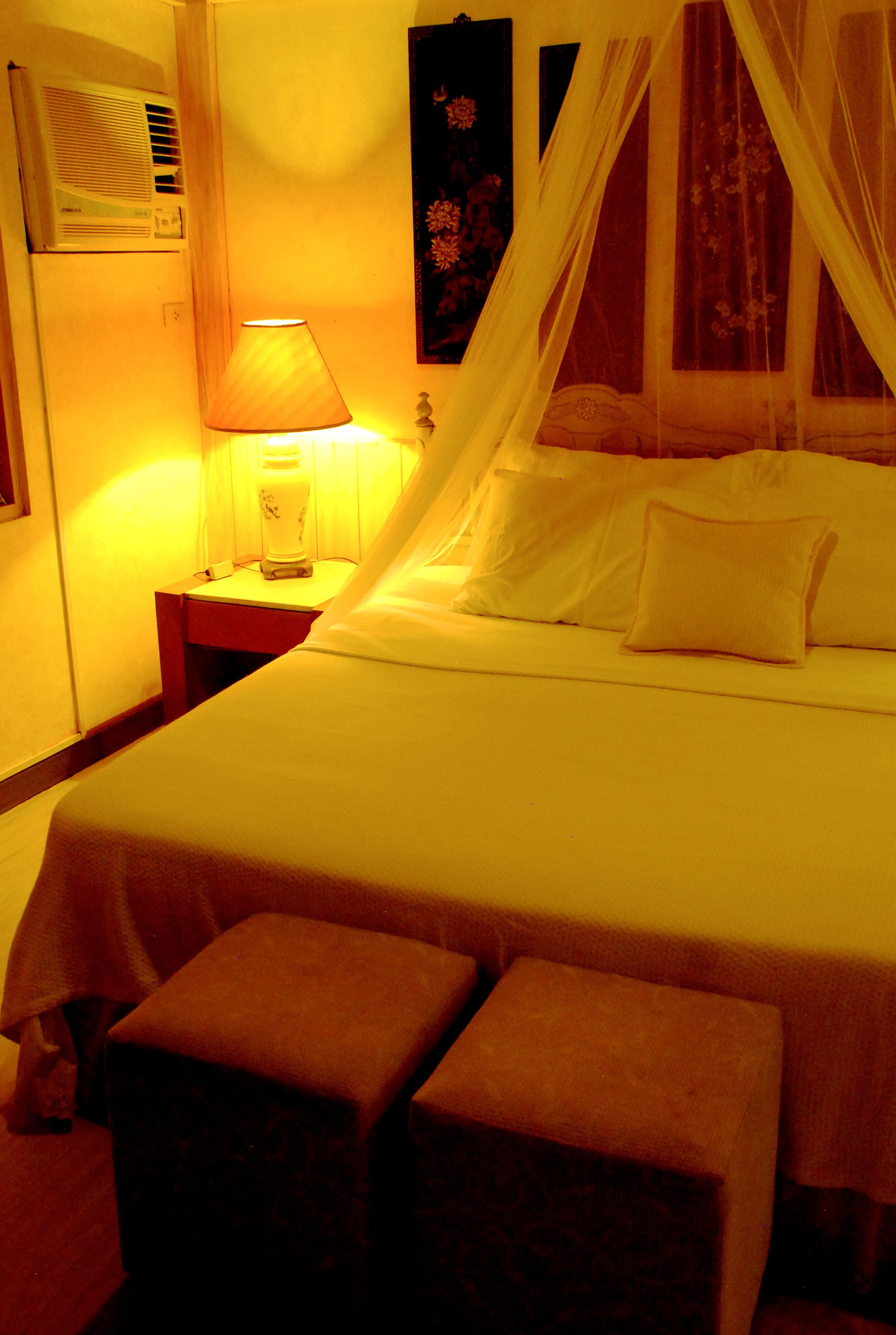 Una habitación de hotel con cama, mesita de noche, lámpara y ventana con cortinas.