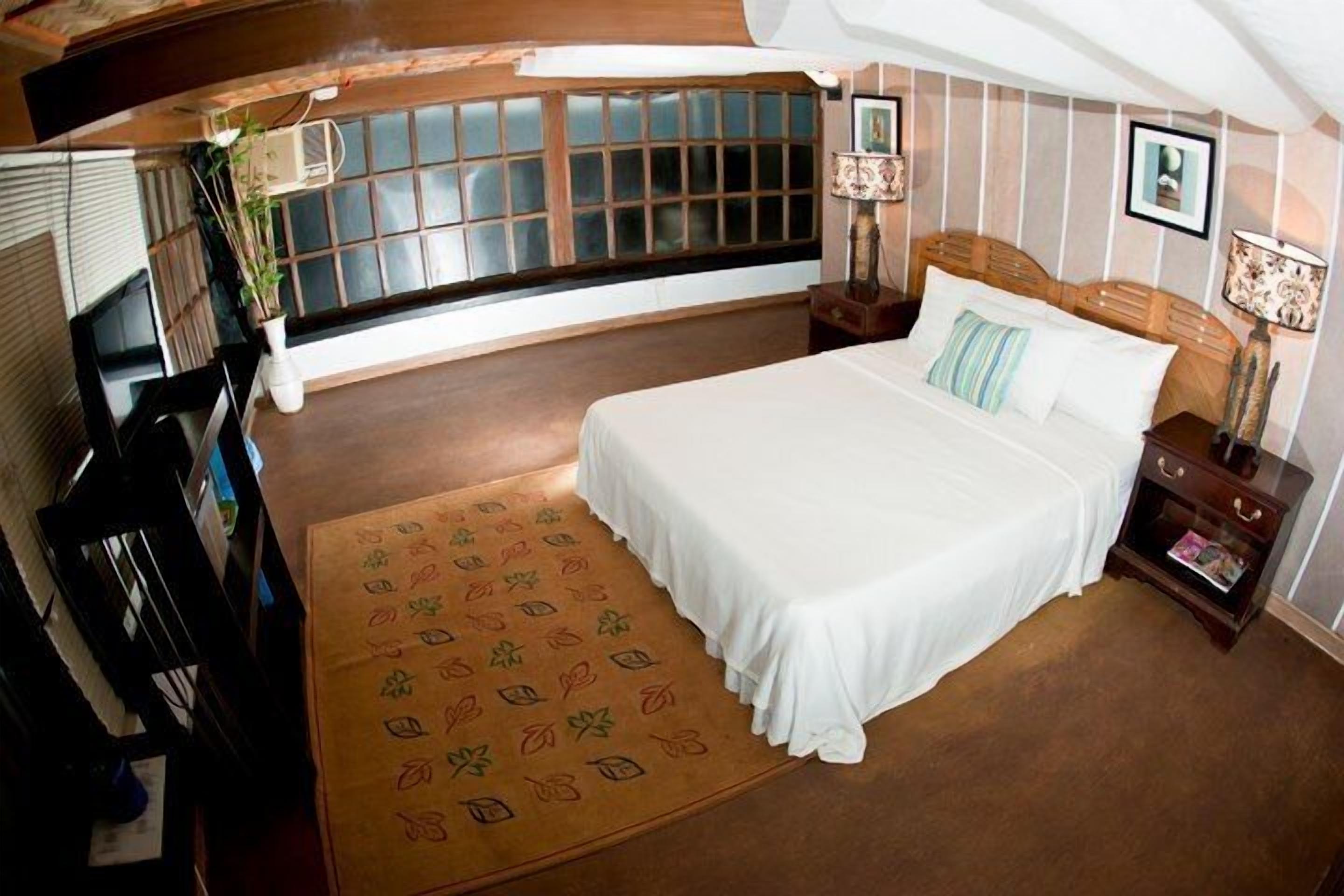 Une chambre à coucher avec un grand lit, une fenêtre avec des stores, une table de chevet avec une lampe et un tapis décoratif.