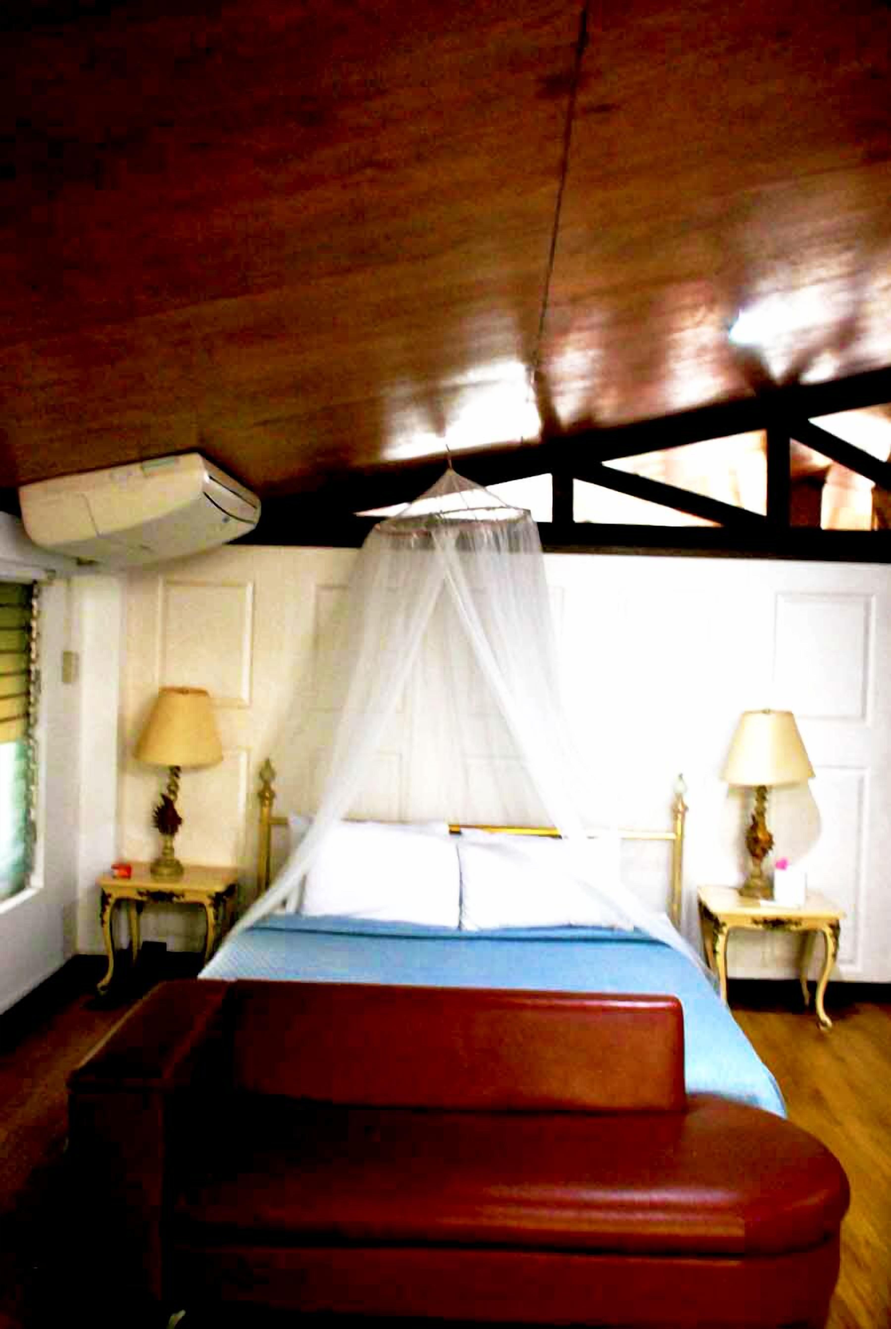 A room with a bed under a wooden ceiling, a window with blinds, and two bedside tables with lamps.