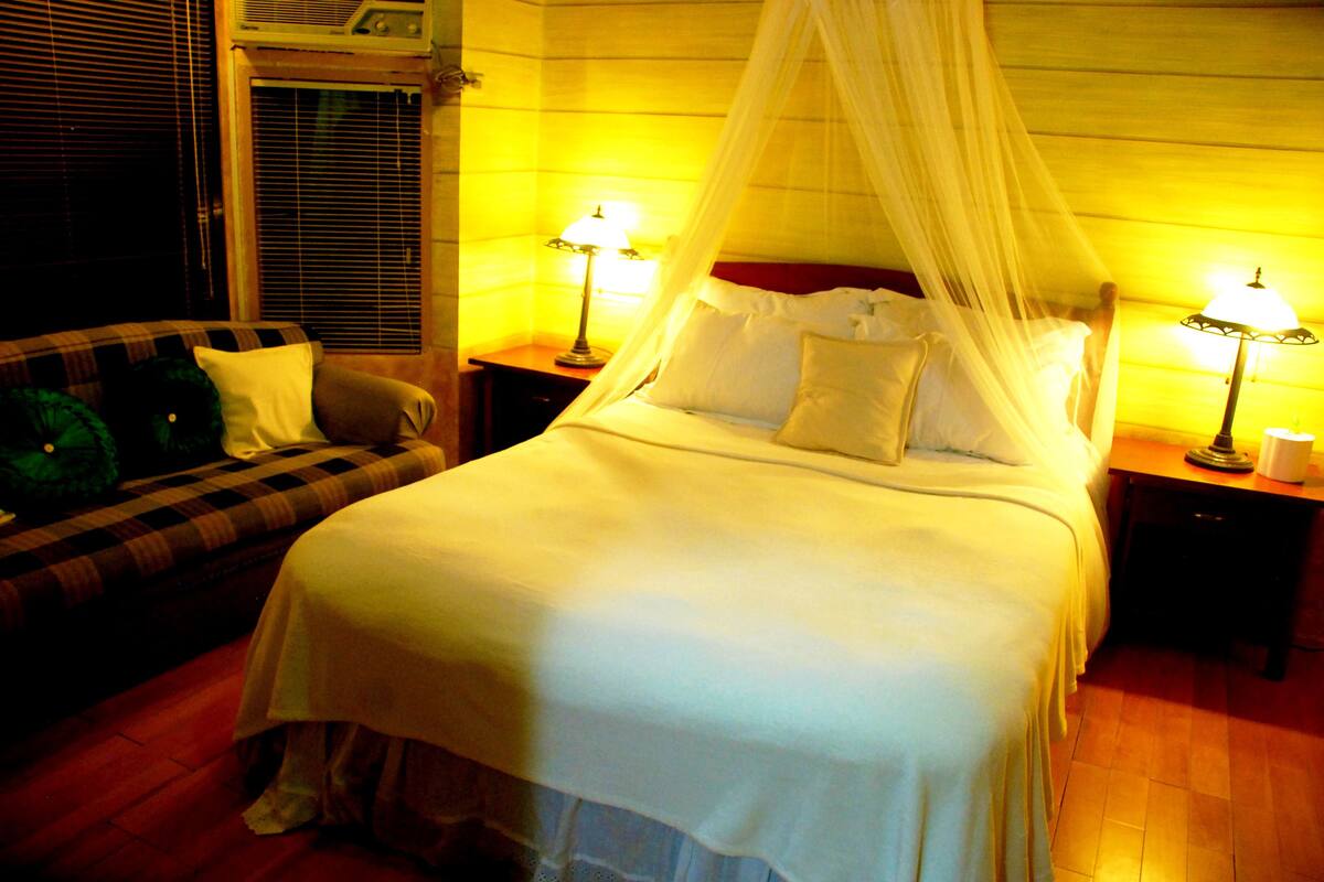 A bed with a canopy, two bedside lamps, a checkered sofa, and a window with blinds.