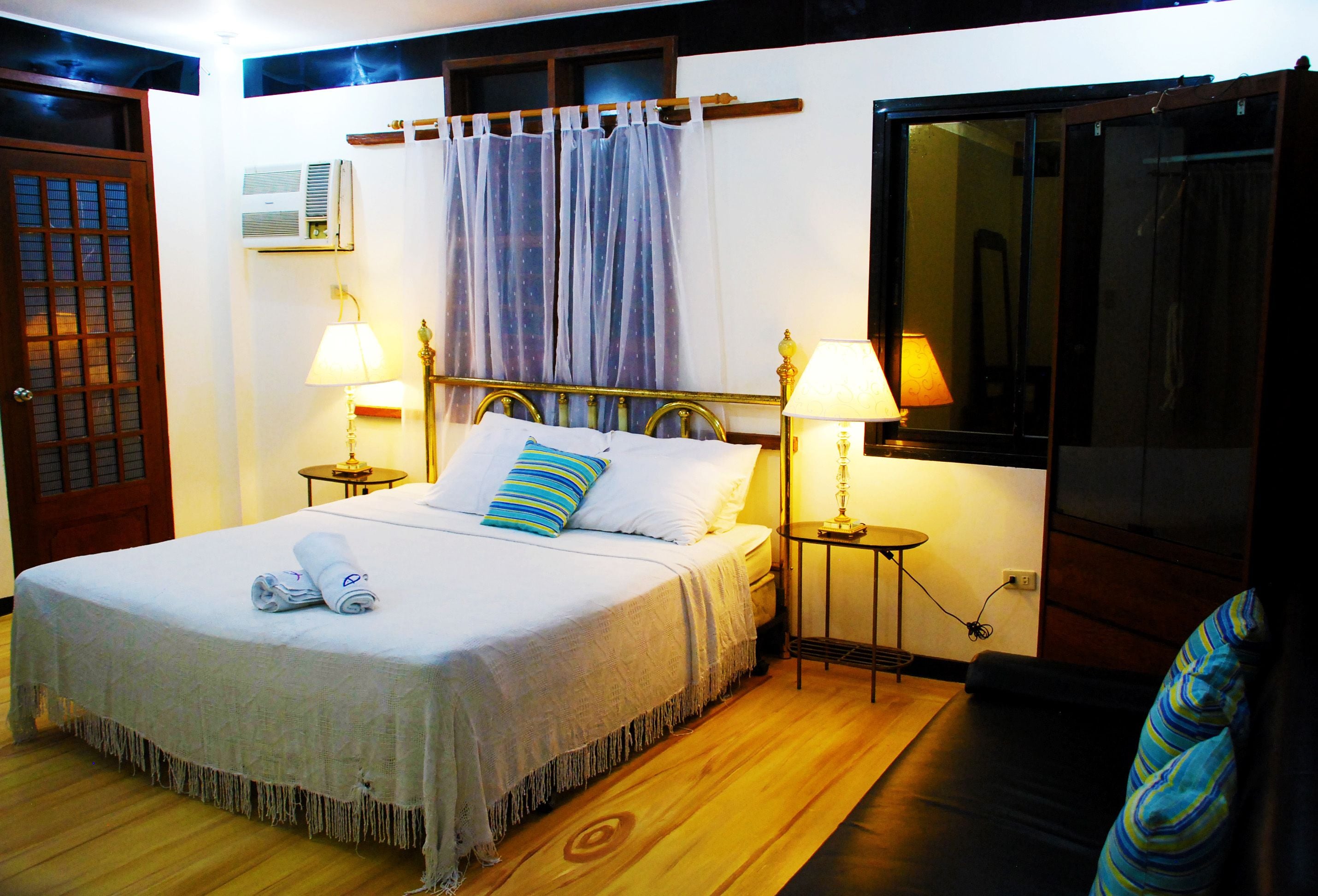 A neatly made bed with a white bedspread and two pillows, two bedside lamps, a window with curtains, and a wooden floor.