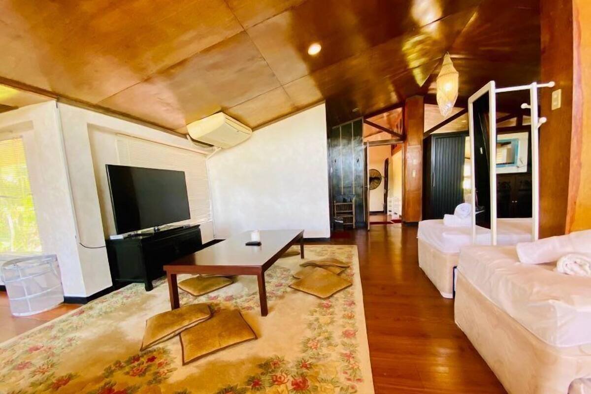 A spacious living area with a flat-screen TV, a wooden coffee table, and a floral rug. There is a white sofa and a small table with towels.