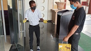 Un hombre con camisa blanca y pantalones negros limpia un ventanal con un cubo amarillo. Otro hombre con camisa negra y jeans permanece cerca sosteniendo un cubo amarillo con un personaje de dibujos animados.