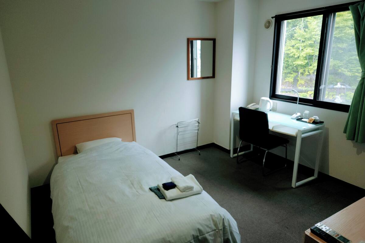 Ein Hotelzimmer mit einem Einzelbett, einem Schreibtisch, einem Stuhl, einem Fenster mit Vorhängen und einem kleinen Heizkörper.