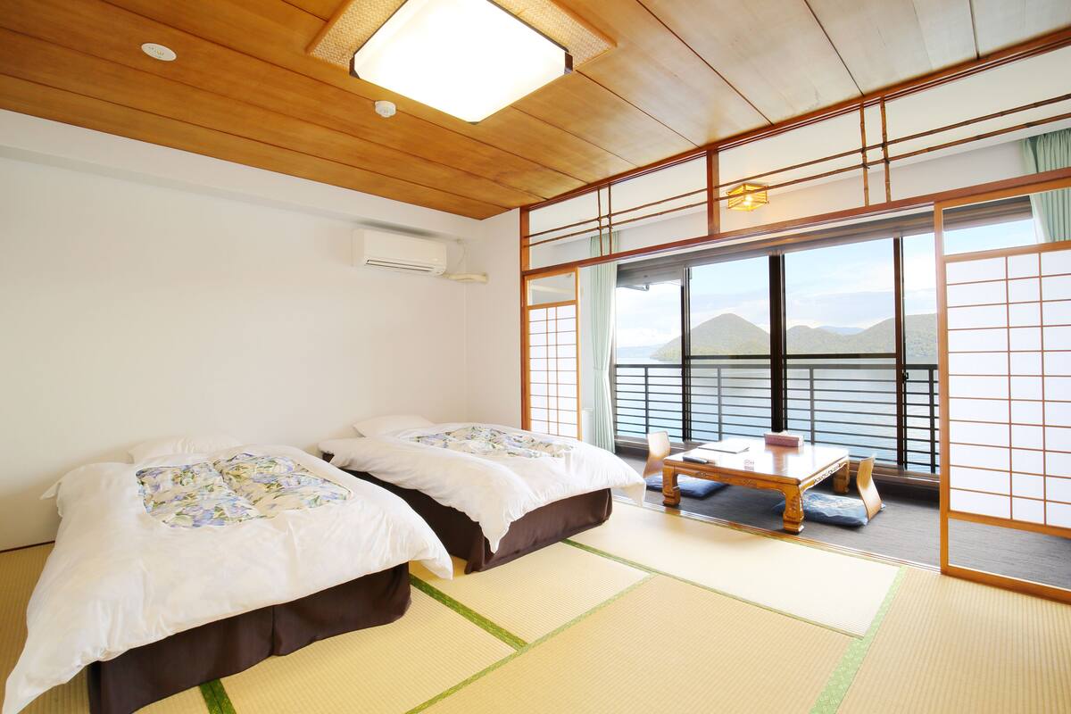 Ein traditioneller japanischer Raum mit Tatami-Boden, einem Bett mit Blumenmuster und einem kleinen Tisch, mit Blick auf Berge durch große Fenster.