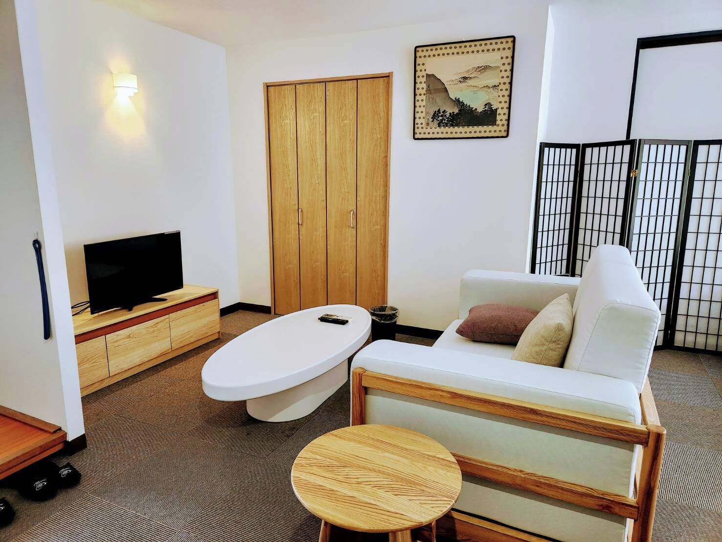A modern living room with a flat-screen TV on a wooden stand, a white sofa with cushions, a round wooden table, and a wooden wardrobe.