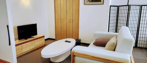 Una sala de estar moderna con un televisor de pantalla plana sobre un soporte de madera, un sofá blanco con cojines, una mesa redonda de madera y un armario de madera.