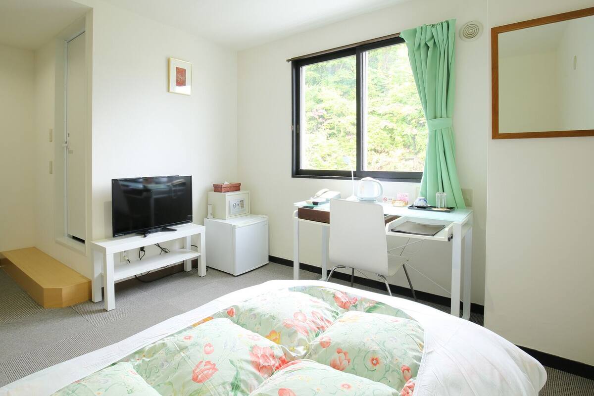 Ein Hotelzimmer mit Bett, Schreibtisch mit Stuhl, Fernseher, Waschmaschine und einem Fenster mit Vorhängen.
