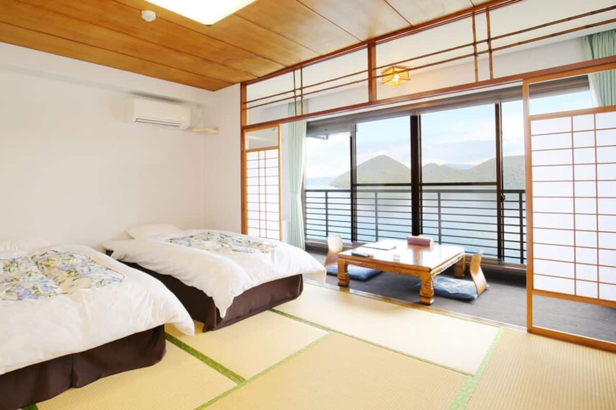 Ein traditioneller japanischer Raum mit Tatami-Böden, zwei Betten mit floralem Bettzeug, einem kleinen Tisch und Blick auf Berge durch große Schiebetüren.