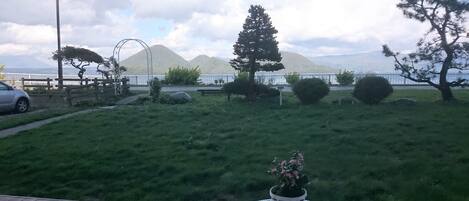 Una terraza con una mesa y sillas blancas, una planta en maceta y vistas a un área verde con árboles y una montaña distante.