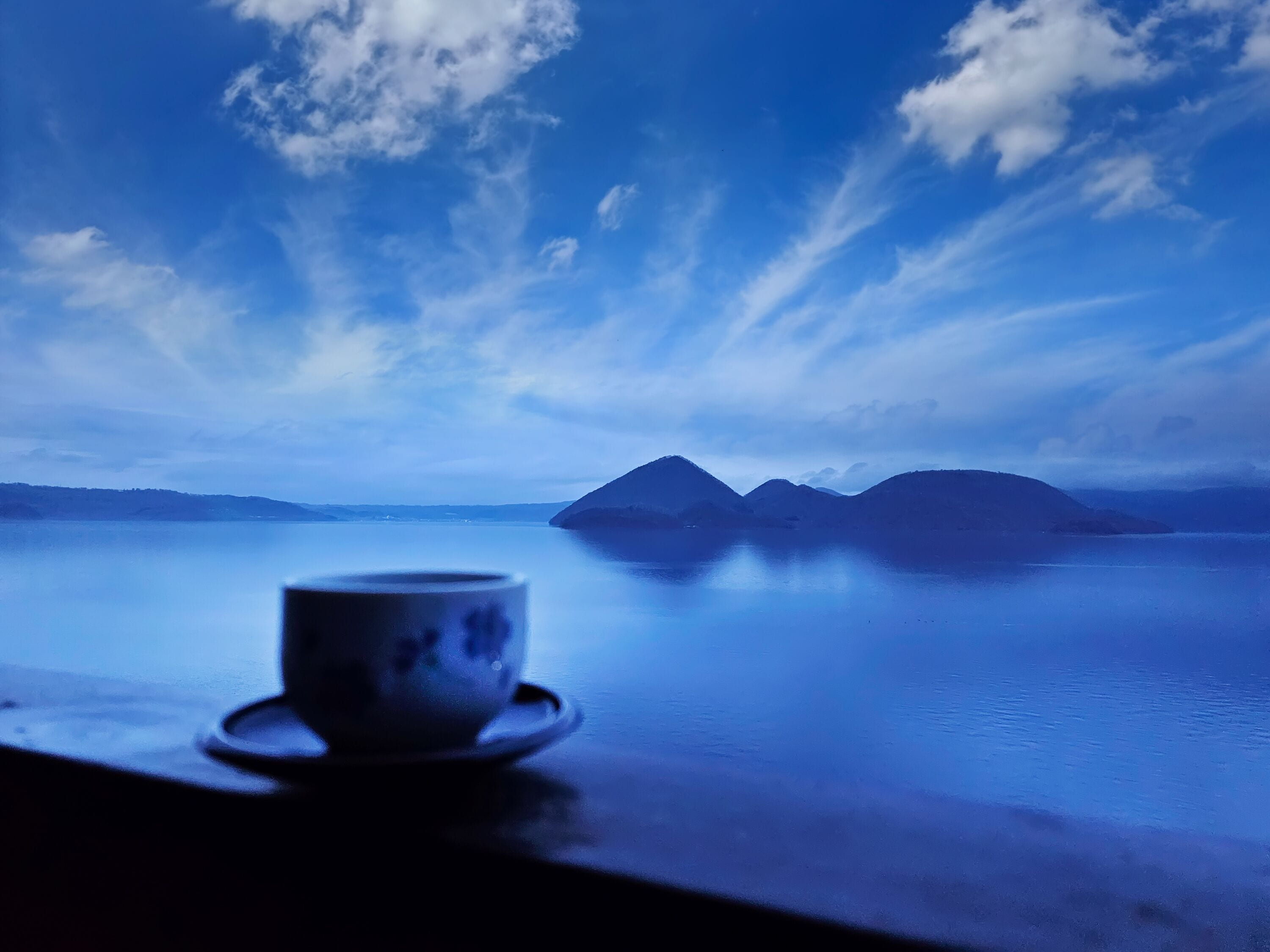 A cup on a table with a view of mountains and a body of water under a blue sky.