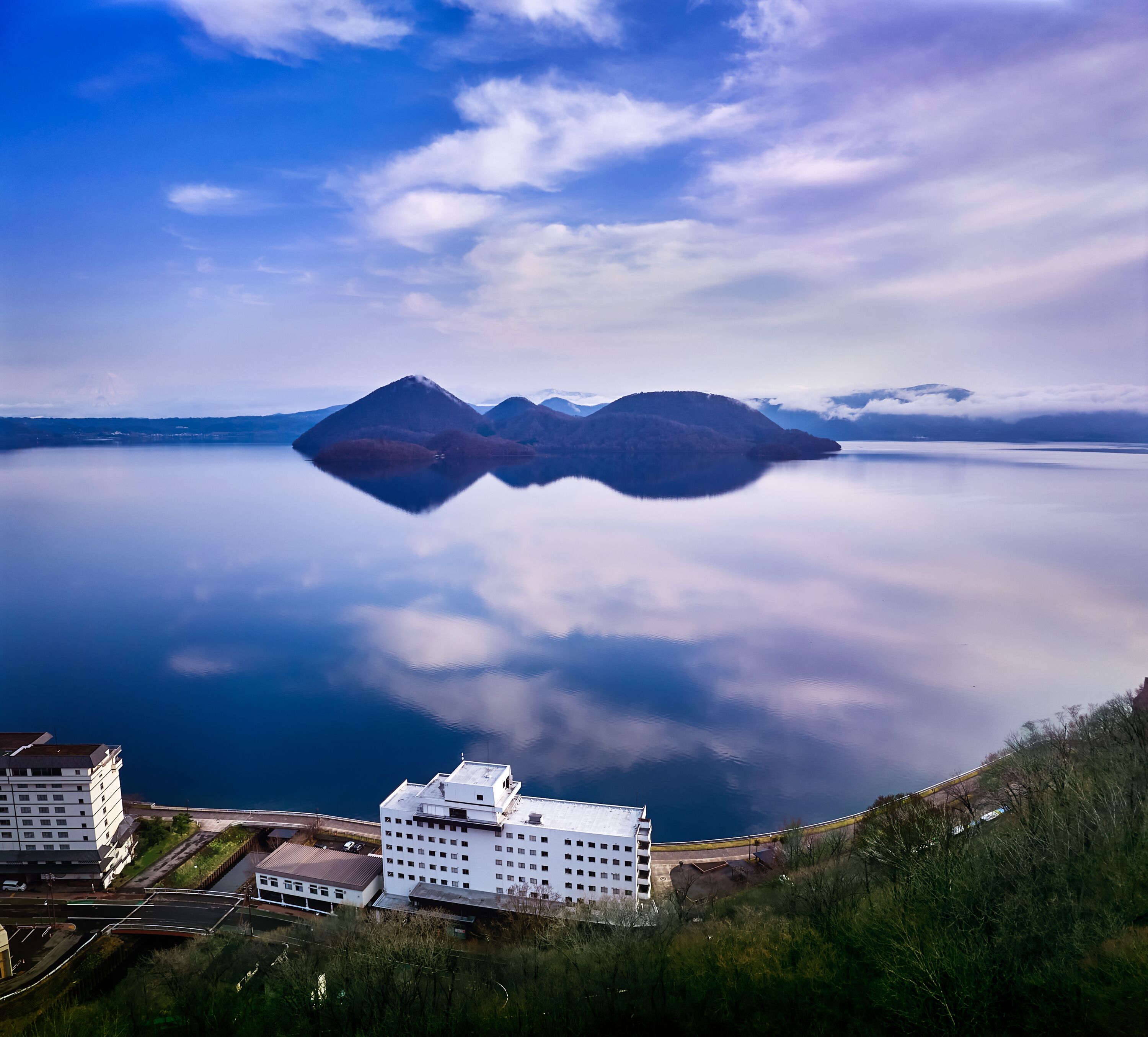 A view of a calm lake with mountains in the distance and a building with multiple floors in the foreground.
